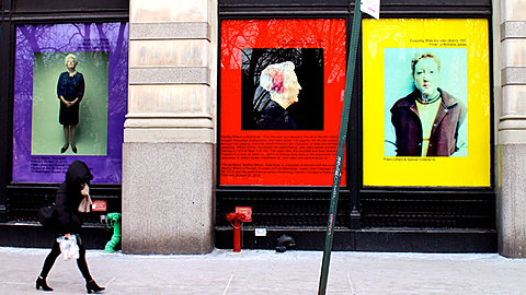 A person walks in front of three large window displays, one purple, one red, one yellow. Each shows the image of a person along with some text.