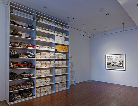 The is a corner of a room, the floor is wooden and empty. The walls are white and the lighting is dim. There is a black and white framed photograph on the right wall that has the only light turned on shining on it. On the left wall there is a white floor to ceiling shelving unit filled with clothing and boxes of printed photographs. To the right of the shelving unit this is a white ladder leaning against the wall.