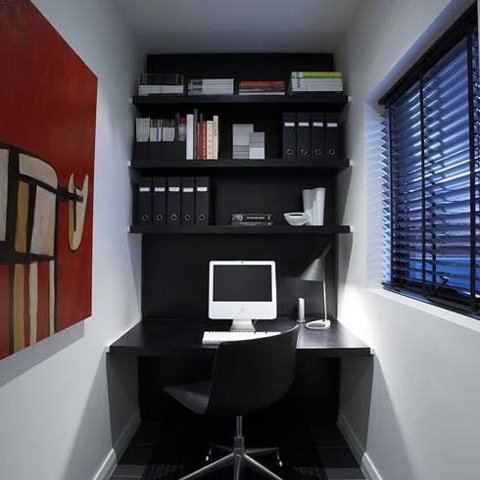 Interior image of installation that resembles the interior of an office. On a black desk sits a computer desk top. Above features a shelving unit with books and other office supplies such as binders, folders. On the left wall is a red painting with a red background and abstract image of an animal outline. On the right is a window with black blinds, open for light to come in. 