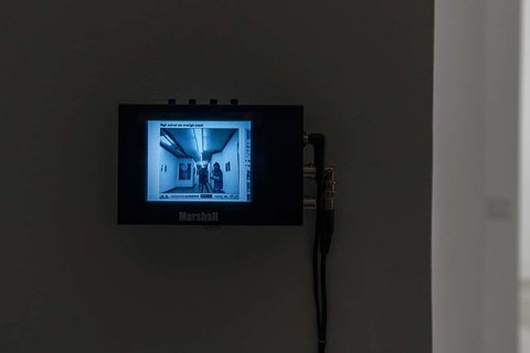 Close-up installation view of a television monitor mounted on the wall with a still from the video, although it is illegible from the camera viewpoint. 