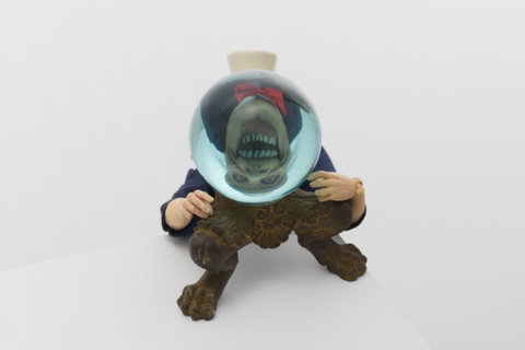 Close-up image of a figurine holding an orb in front of its face. The orb reflects the face upside down and reveals a terrifying, open-mouthed monstrous figure with deep set eyes. The figure wears a white top hat and has stringy, grey hair. It's arms are wrapped around the orb which sits upon a brass claw foot pedestal. 