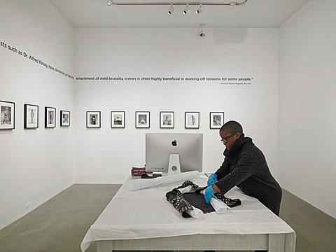 There is a white walled room with concrete floors. In the middle of the room is a table with a sheet of fabric laid over it and a desktop on it. There is also a student working on unrolling prints. The wall to the back that it perpendicular to the table has a line of framed black and white photos that continue onto the adjoined perpendicular left wall. Above the photographs is a quote that also follows from the back wall onto the left adjoined perpendicular wall. 