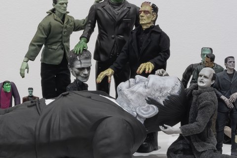 Image of a sculpture in the exhibition featuring several figurines crowded around a larger figurine who seems to have fainted. The figurines include several variations of Frankenstein crowding around a large version of Frankenstein. 