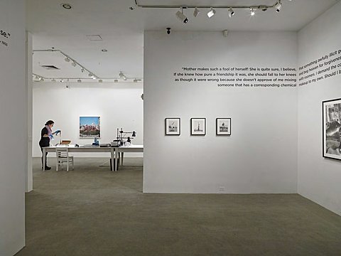 There is an open floor plan that has a room with half length walls that show the room behind it. In the more forward room the concrete floor is empty and the two visible white walls feature black and white framed photographs. There are three small photographs on the wall parallel to the camera, and one larger photograph on the right perpendicular wall. Above the photographs isa quote the spans both of the walls. In the room behind this one is a table with a student working to develop and print photographs. On the wall parallel to the camera in this room there is a colorful photo that features a vibrant blue. 