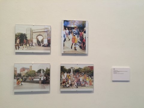 Close-up installation view of four works in the exhibition arranged two on top of two. The works are photographs from various spots in Washington Square park. In the top left image, there is a line of people, presumably a fashion show, walking from the arch in Washington Square Park. The top right image, features a single figure modeling clothes with black and white stripes and blocks of warm, orange color. The bottom left image features a still shot of the fountain in Washington Square Park with many people surrounding it, going about their daily lives. In the bottom right image is a group shot of the models dressed in black, white, and warm colors in Washington Square park. Beside the images, on the right is a plaque with the title, however it is illegible from the photograph.  