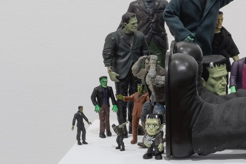 Image of a sculpture in the exhibition featuring several figurines crowded around a larger figurine who seems to have fainted. The figurines include several variations of Frankenstein crowding around a large version of Frankenstein. 