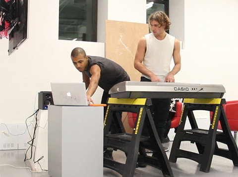 Installation view featuring to figures engaging in a makeshift DJ booth. The figure on the right the leans over to use a Macbook sitting atop a white pedestal. The figure wears a black tee shirt and has a close-cropped haircut.The figure on the right wears a white muscle tank top and places both hands on a keyboard or disc jockey table that is balanced on two construction supports.  