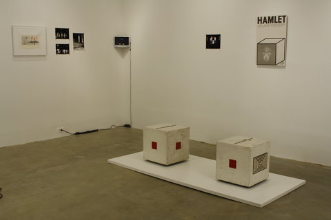 Gallery installation view of the exhibition. A white, rectangle platform is on the ground. On top of the white rectangle are two white cubes. On the sides of the white cubes, there are small red squares on the sides of the cubes. Behind the installation on the floor, there are seven images on two walls. On the left, there is a white frame with a sketch. Next to sketch are two black squares with white figures, the photo is not legible from the photo provided. Adjacent to these two images is one image with similar content of black image with white figures. In the corner, there is a small monitor with nothing on the screen. On the right hand wall, there is a black square. Adjacent to the black square is a poster with the word HAMLET over a three-dimensional drawing of the outline of a cube.   