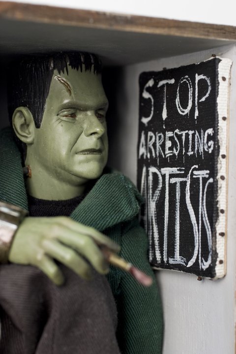 Close-up image of a figurine sculpture featuring a Frankenstein character figurine with green skin, black hair, and a green jacket. The figure is next to a black sign with white writing that says, "STOP ARRESTING ARTISTS." The figure holds a paintbrush as if it recently finished painting the sign. 