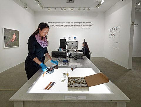  A student looks at strips of photographic film on top of a Lightbox as she prepares them to be developed. Perpendicular to the table she is on is a wall with a quote printed on it. On the left wall parallel to the Lightbox table is a colored photograph of a man laying in foliage. On the right parallel wall to the Lightbox table there are clotheslines and pins placed to hold drying freshly developed photos. 