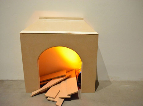 Close-up image of installation featuring a hollow, wood box with a orange light and wood planks inside. The planks are falling out of the arch shaped hole and resemble a simplified, fake fireplace.  