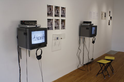Gallery installation view of the exhibition. On the wall from left to right is a television featuring a white still image. Hanging on the television is a pair of black headphones. Next to the television is a collection of six images grouped two vertically and three horizontally. The images consist of a figure in front of a table in six different poses. Adjacent to this group of images is another television set. This one is illegible from the viewpoint. There is a pair of black headphones hanging from this monitor as well. On the floor are two stools in front of the television sets.  