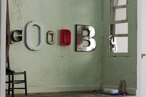 Close-up image of letters hung on a green wall of a model room. The letters, G, O, O, D, B, and Y are hung on the wall in various fonts. The letter 'Y' is being used to prop open the fake, model window and other letters are scattered on the floor. On the right there is a chair with a telephone on it. 