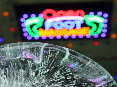 Installation view of the exhibition featuring a very up-close shot of a glass orb. Behind the orb is a blurry neon sign that says "open" in red neon lights, and below that "foot" in blue neon lights. On either end of the sign are green outlines of a foot. 