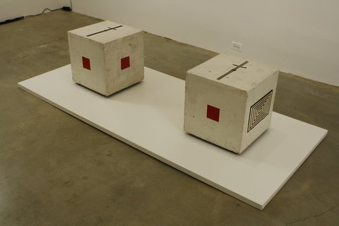 Gallery installation view of the exhibition. A white, rectangle platform is on the ground. On top of the white rectangle are two white cubes. On the sides of the white cubes, there are small red squares on the sides of the cubes.  