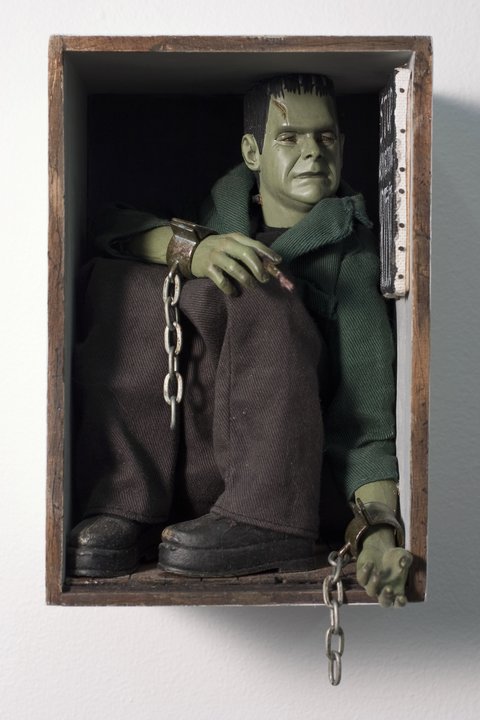 Close-up image of a Frankenstein character figuring crouched in a small box. The figurine has green skin, a green shirt, black hair, black boots, and brown pants. It is holing a paintbrush in it's right hand. 