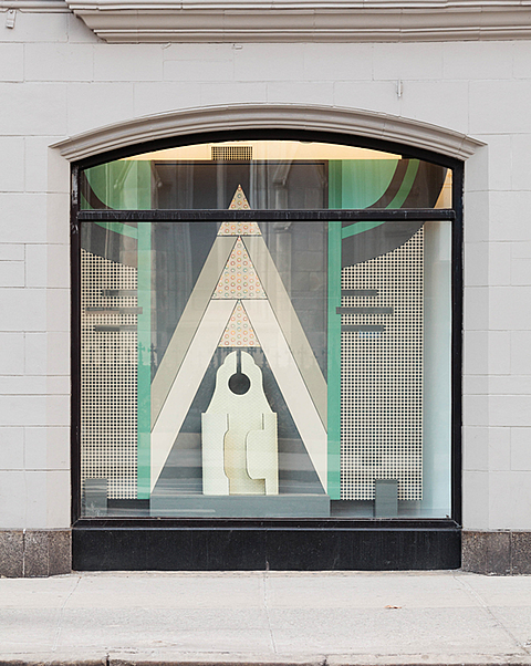 The window is broken into different geometric cut out shapes. The two outer thirds of the window are filled with a rectangular mesh like board that is lined with teal and broken up by alternating teal and black circle quarters. In the middle of the window is a triangle that is broken into three triangles that get subsequently smaller. The tallest is a light brown, the middle is white, and the smallest is black. The top tips of each of the triangles has a smaller triangle that is tan with colored circles on it. In front of the smallest black triangle is a rounded paddle-like cream colored cutout. 