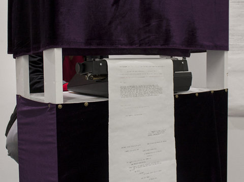 Close-up image of a typewriter with a long typed page emerging from it. On the top and bottom of the typewriter are purple boxes supported by white stilts. 