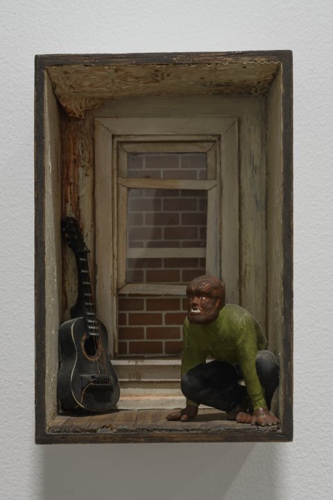 Image of a sculpture in the exhibition featuring a model version of a wooden hallway. At the end of the hallway, an open-window reveals a red brick wall. In the hallway, crouched on the ground is a werewolf character figurine wearing a green long-sleeve top and dark pants. Opposite the figurine, a dark colored guitar figurine leans against the wall. 