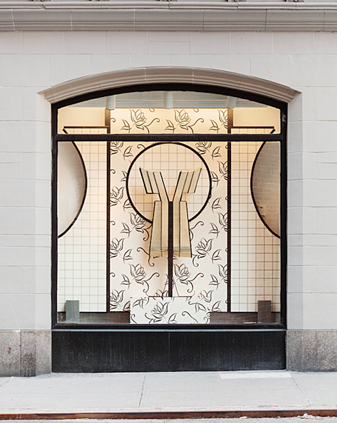  The outer two thirds of the window have a gridded  cream colored rectangular board placed in them with a half circle cut out from their top outer edges. The middle third features a cream colored rectangle with a repeated flower pattern printed on it in gray. In the middle of this middle rectangle is a circle cutout of the gridded rectangle board from the outer two thirds of the window space. In front of the middle patterned cutout is a tan wooden sculpture of the silhouette of a dress with a deep V neck, swooped shoulder line and pleated skirt. 