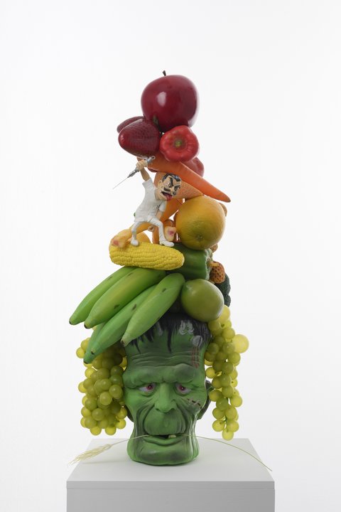Close-up detail image of a sculpture in the exhibition featuring a stack of fruit and vegetables. From top to bottom, the sculpture includes corn, oranges, carrots, bell peppers, and strawberries, arranged in a topsy-turvy tower pattern. A figurine stands on the corn in a white chefs top, white pants, and white boots. The figure has a cartoonish face, with bulging eyes, raised eyebrows, a large red tongue sticking out of its mouth and dark brown, slick backed hair. It wields a large needle in one arm and looks crazily at the fruit as if it is about to attack. Underneath this figurine is a model of fake green bananas and grapes. All of these figures sit atop a large green Frankenstein head that holds a piece of wheat in its mouth. 