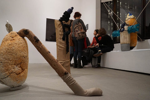 Installation image of visitors frequenting the exhibition space. The visitors are gathered in the background. Pieces from the exhibit sit on the floor in the foreground. 