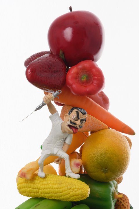 Close-up detail image of a sculpture in the exhibition featuring a stack of fruit and vegetables. From top to bottom, the sculpture includes corn, oranges, carrots, bell peppers, and strawberries, arranged in a topsy-turvy tower pattern. A figurine stands on the corn in a white chefs top, white pants, and white boots. The figure has a cartoonish face, with bulging eyes, raised eyebrows, a large red tongue sticking out of its mouth and dark brown, slick backed hair. It wields a large needle in one arm and looks crazily at the fruit as if it is about to attack. 
