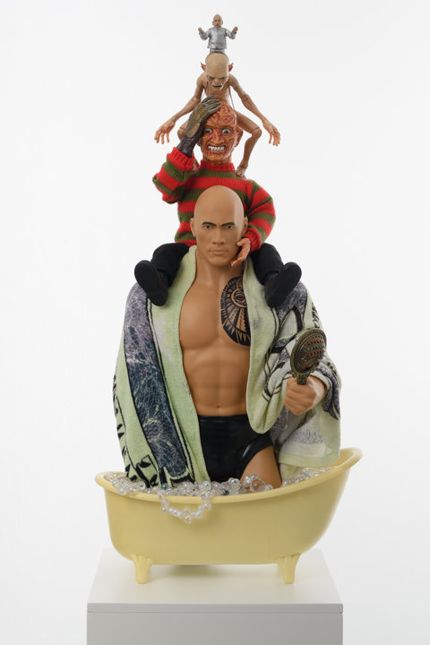 Image of a sculpture in the installation. From bottom to top, a yellowish bathtub filled with iridescent white beads sits atop a white pedestal. In the bath, from the knees up is a muscular figurine with no shirt on. The figurine wears small black shorts, and is wrapped in a towel designed like a one dollar bill, the figurine holds a bedazzled handheld mirror in one hand and stares directly at the viewer with sharp, blue eyes and dark eyebrows. The figuring is bald and has a large, black tattoo on its pectoral. On the shoulders of the figurine sits the black pants and shoes of a Freddy Krueger character figurine. In the classic red striped sweater, the figurine holds its gloved hand up to it's burnt face, showing gritting white teeth. Atop this figurine, sits a Gollum character figurine with its arms outstretched, standing on the shoulders of the figurine below. Atop the Gollum character figurine, is a very small Dr. Evil character figurine in a classic silver suit and bald head. 