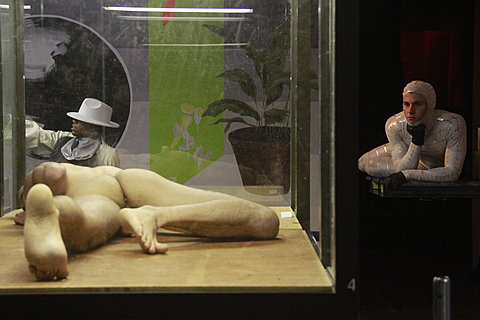 A nude man lying across the floor face down is in a glass display case. Outside the case, in the corner on the right, is man in a white jumpsuit on a pedestal. His head is resting beneath his chin.