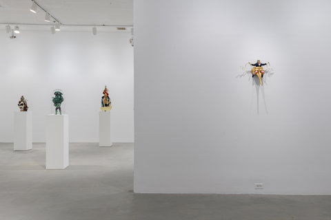 Image of four works in the exhibition with the view of three sculptures on pedestals scattered throughout the background. In the foreground, on the wall hangs a monstrous Uncle Sam figurine sitting on a gold mask with a long nose. The figurines red and white striped legs come through the eyeholes of the mask. The figurine's arms are outstretched and hold a coiling, gold ribbon in each on. The figurine wears a navy blue coat, with gold buttons, and a white undershirt and red bowtie. With deep set eyes and scraggly grey hair emerging from it's white top hat, the figurine is posed open-mouthed at the audience with a crazed look upon it's face. 