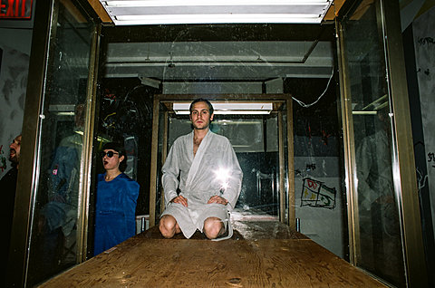 A man in a grey robe is sitting on his knees on a wood floor within a glass chamber.