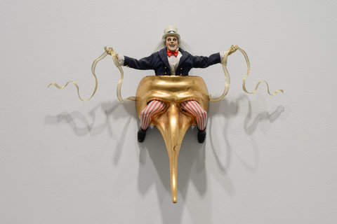 Image of a sculpture in the exhibition featuring a monstrous Uncle Sam figurine sitting on a gold mask with a long nose. The figurines red and white striped legs come through the eyeholes of the mask. The figurine's arms are outstretched and hold a coiling, gold ribbon in each on. The figurine wears a navy blue coat, with gold buttons, and a white undershirt and red bowtie. With deep set eyes and scraggly grey hair emerging from it's white top hat, the figurine is posed open-mouthed at the audience with a crazed look upon it's face. 