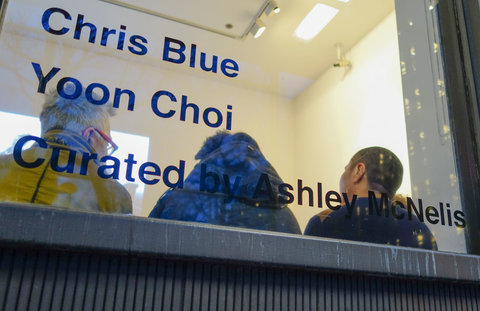 Image of the exhibition from the outside window featuring a room with three people seated against the window. On the window, the words "CHRIS BLUE, YOON CHOI, CURATED BY ASHLEY MCNELIS" are on the window in blue vinyl wall text. 