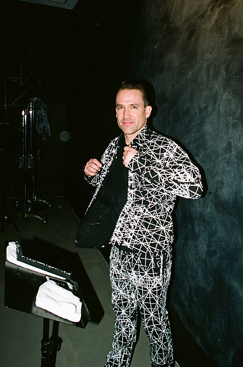 A maA fair toned man with slicked back hair is wearing a black and white patterned suit. He has a slight smile and appears to be opening the jacket. n in a white suit with green spots on it lays face up on a white circle on the floor.