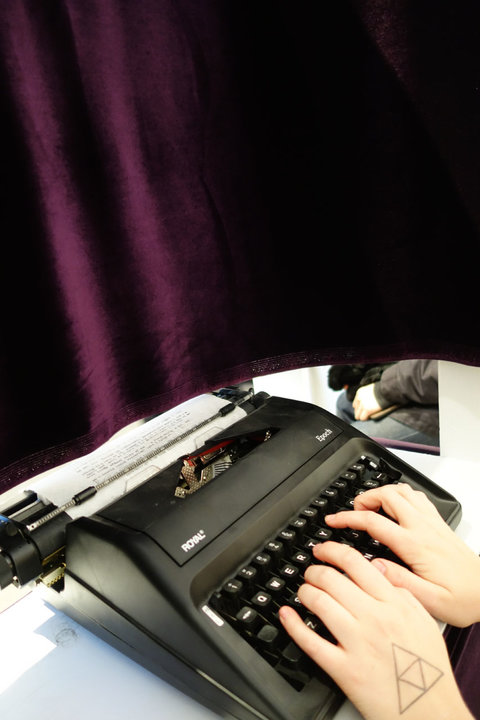 Close-up image of two hands typing on a type writer. In front of the type writer, a burgundy-purple velvet curtain hangs and obstructs further viewpoints. 