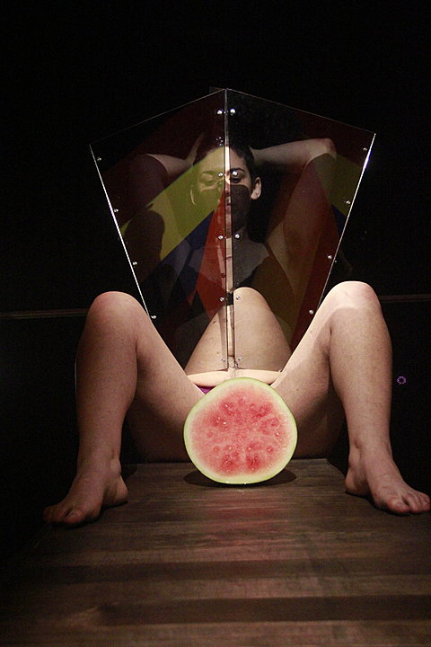 In a dim lit room a partially nude woman sits with her legs spread open. In-between her legs is a watermelon cut in half. There is a glass barrier placed by her lower abdomen that sections off her legs from her upper body.