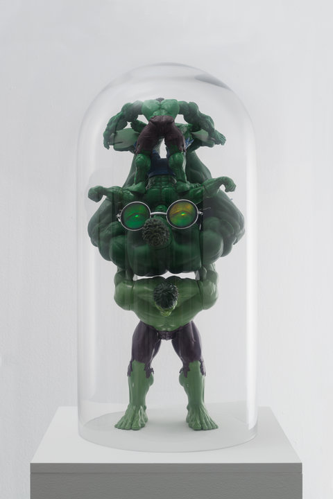Image a sculpture in the exhibition featuring a conglomeration of multiple figurines of "The Incredible Hulk' stacked upon one another inside a glass half-cylinder.