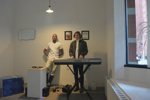 Two figures play keyboard in front of two images on a white wall. They are dressed in white tee shirts. 