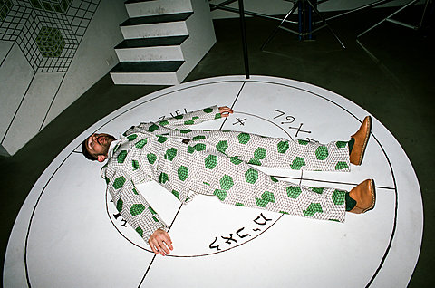 A man in a white suit with green spots on it lays face up on a white circle on the floor.