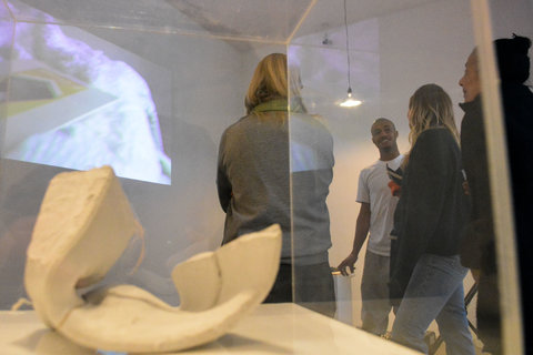 Close-up image that features the inside of a glass container featuring a white sculpture inside. Through the glass, there are various figures standing in the gallery, visitors watching a video projected onto a wall. 