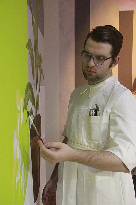 A man with fair complexion, dark hair, glasses, and short beard is holding a paint brush. He is wearing a white apron with more brushes in the upper right pocket. He is using green paint to create leaves of a plant on the wall to his left. 