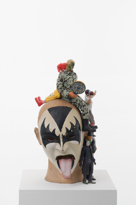 Image of a sculpture in the exhibition featuring the head of a Gene Simmons from KISS character, with a bald head and white painted face with black lips and a black triangle on the forehead and underneath the eyes. Other smaller character figurines surround the head. The red shoes, red hair, and yellow outfit of Ronald McDonald are seated on the left of the head back to back with a army character dressed in camo, with a camo painted face in green, grey, and brown. Next to the head, on the right side,  whose tongue is sticking out of its mouth, is a Joker character figurine complete with a long black jacket and boots. The Joker has a white painted face, red lips, and green hair. On top of the Joker figurine, facing away from the viewer is a football player figurine dressed in full gear. The figurine's arm is holding a football, pulled back and ready to launch the football.   