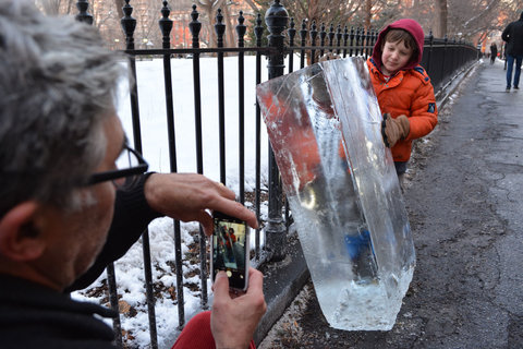 Close-up image of an older male figure taking a photograph of a younger figure holding a very large block of ice. The camera angle is positioned so that the figure taking the photo is on the left side of the image, out of focus, and the other figure, holding the ice in a red puffer jacket, is in the back right. 