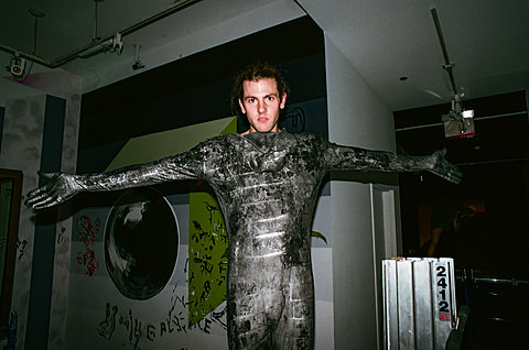 A photograph of a man wearing a sliver and black long-sleeved jumpsuit. The jumpsuit covers his hands with gloves. His arms are extended horizontally.   