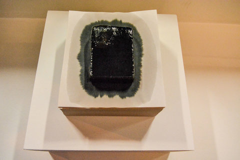 Image of what seems to be an ice cube of black water, melting onto a piece of white paper, on a wooden pedestal. The image is taken from above. 