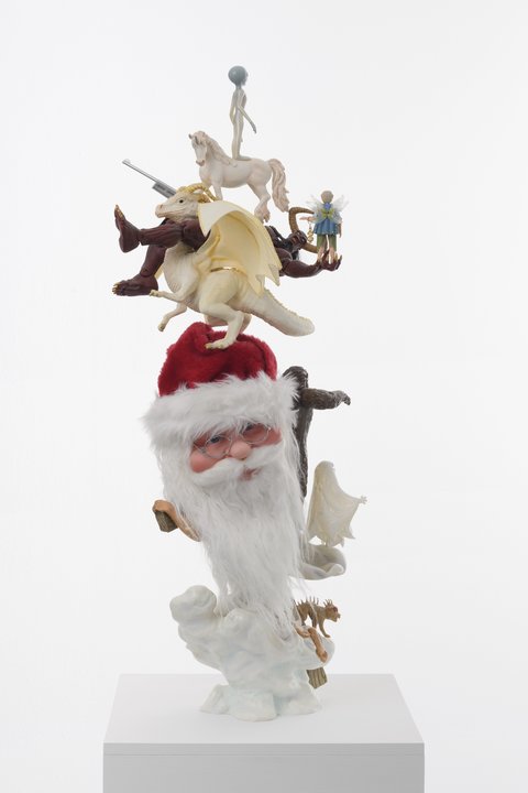 Close-up sculpture in the exhibition featuring a stack of figurines. From top to bottom, the figurines are a small white, shiny alien with a large balloon head and dark teardrop eyes, standing atop a white unicorn, which is standing atop a dark red, satanic figure with fiery eyes and massive horns. The figure is laying flat and its legs are on the shoulders of a white dinosaur-like figure with big yellow horns and big wings. The Satanic figure's palms are outstretched and in its palm holds a small nymph-fairy figure. The dinosaur stands atop a Santa Claus with a classic red hat, long white beard, and wire-rimmed oval glasses. On the back of Santa's head is attached a bigfoot figurine and a small white sheet ghost figurine. At the base of these figurines a Jesus holding a big wooden cross in a white toga. On the bottom of the toga, is a small red-eyed reptilian creature figurine. The very bottom base appears to be white, cloud-like shapes. 