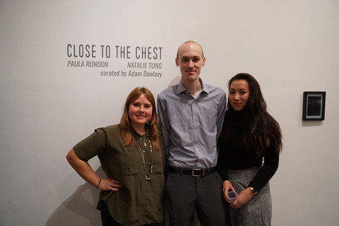 Image of three visitors standing in front of vinyl wall text in the exhibition. The wall text states, "CLOSE TO THE CHEST, PAULA RONDON, NATALIE TUNG, CURATED BY ADAM DUNLAVY."