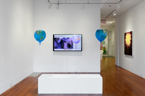 Installation view of a works in the exhibition featuring a white block bench in front of a TV monitor that is mounted to the wall. On either side of the TV monitor are two blue balloons hovering about 5 feet off the ground. Off to the right, an abstract painting of purple and yellow leads off into the hallway. 