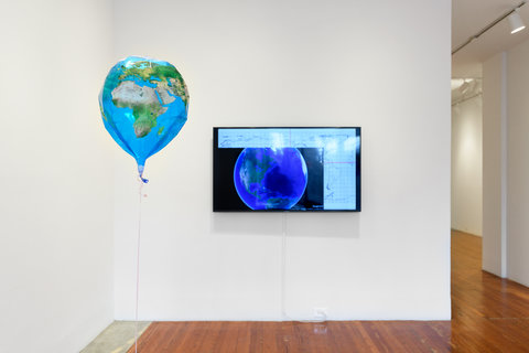 Installation image of a section of the exhibition including a blue earth themed balloon hanging in front of a tv monitor in the background.