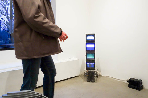Installation view of a figure walking through the exhibition. Only the bottom half of the figure is visible. In the back corner, several small television monitor screens are stacked on top of one another. All four of them are balancing on a clear box with a pair of shoes inside. On the right side of the stack of monitors, a black box for powering the monitors sits on the cement gallery floor. 
